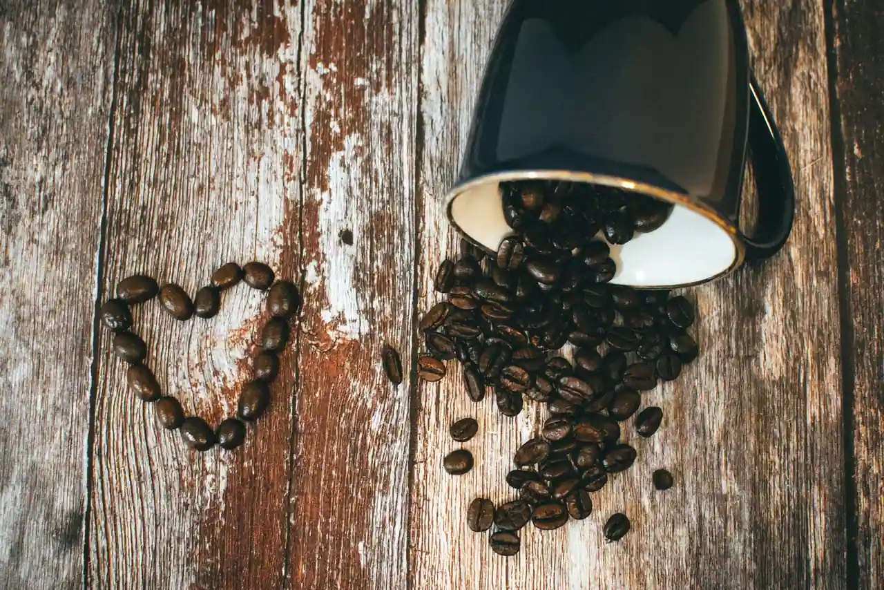 Mug en céramique noire renversé d'où s'échappent des grains de café torréfiés sur un plateau en bois