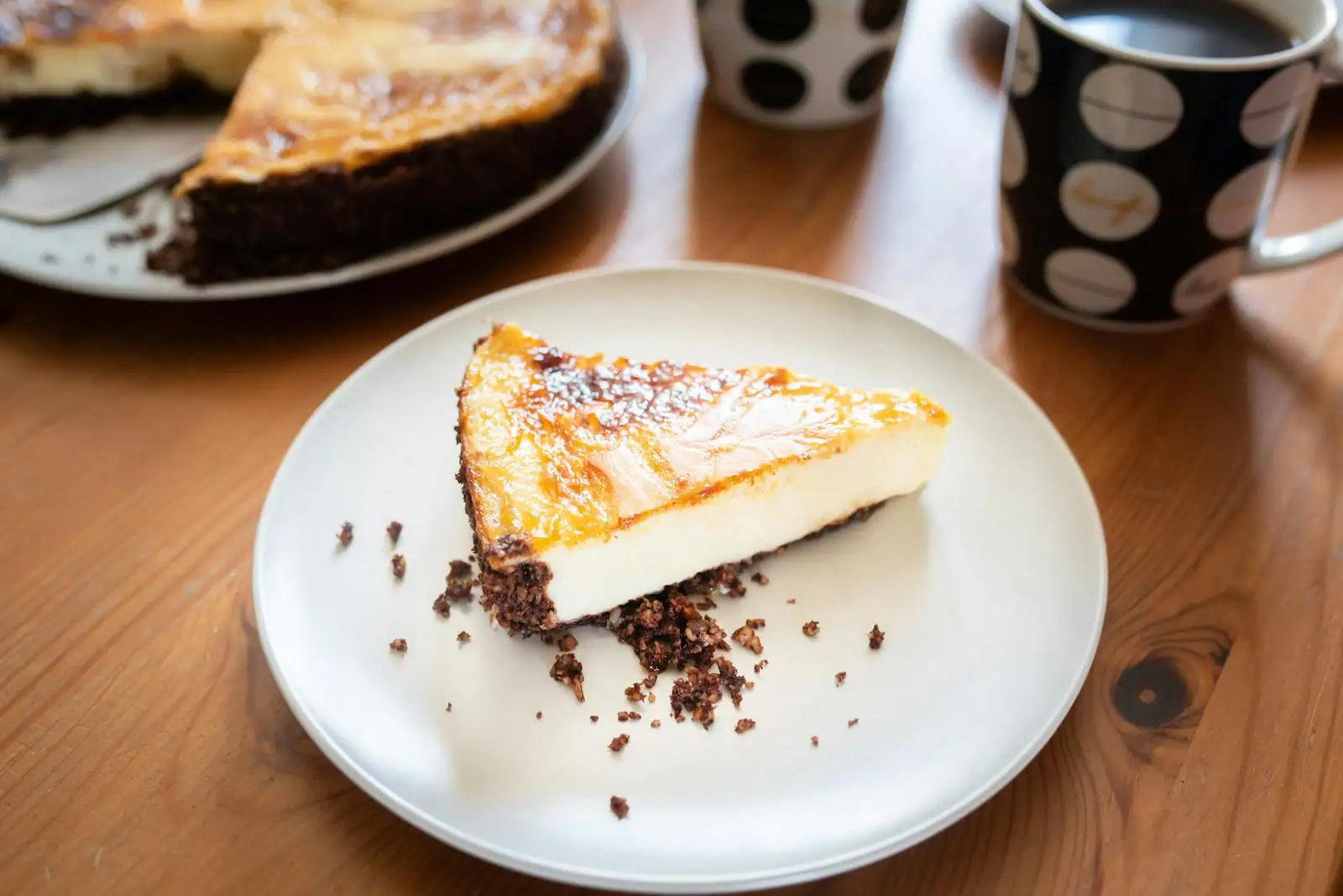 Part de cheesecake doré sur une assiette blanche, deux tasses de café à pois sur une table en bois