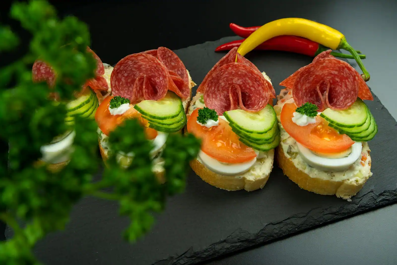 Petites tartines apéritives garnies de salami, concombre, tomate et œuf, dressées sur une ardoise noire