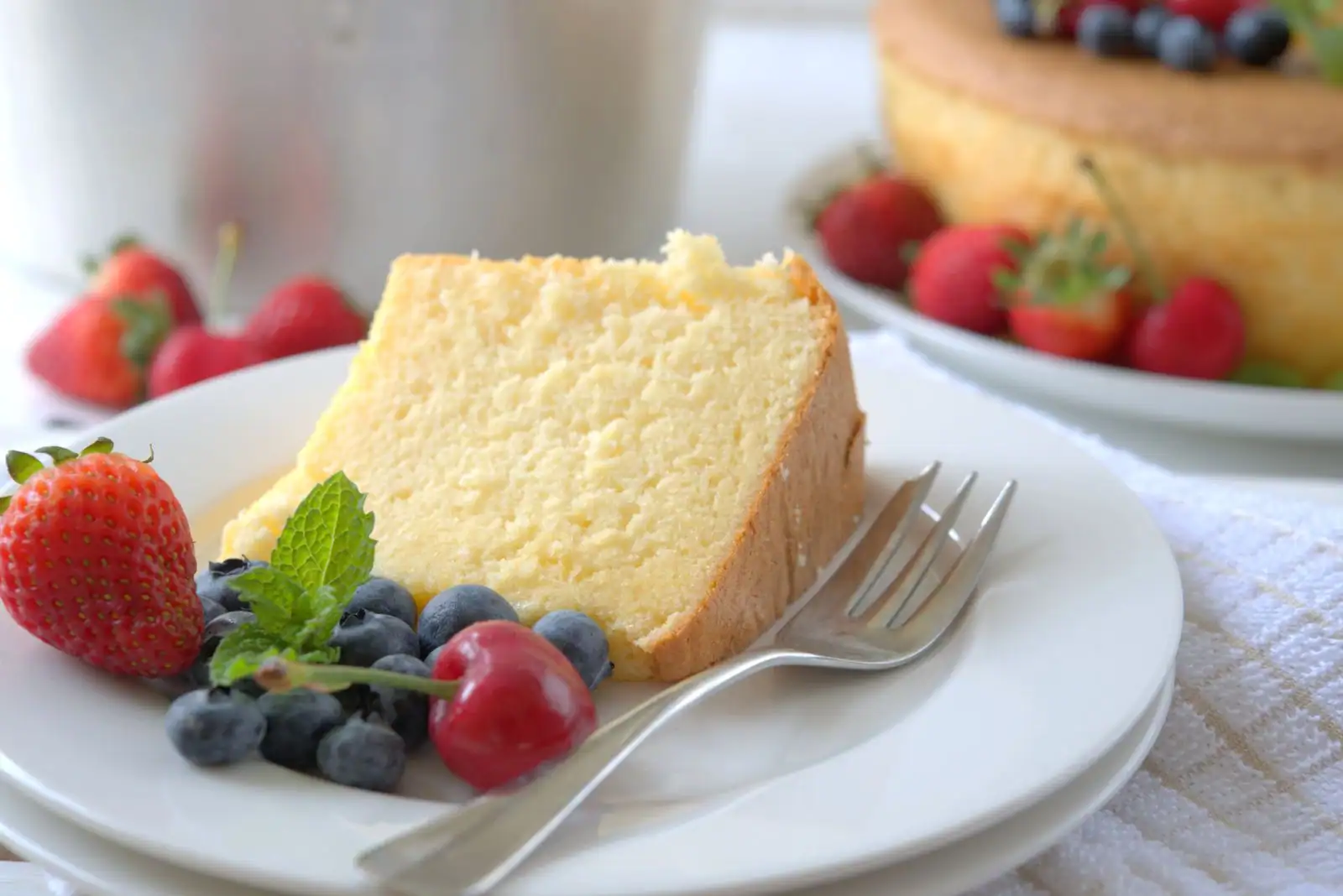 Part de gâteau au yaourt doré et moelleux dans une assiette, accompagnée de fruits frais, gâteau entier en fond