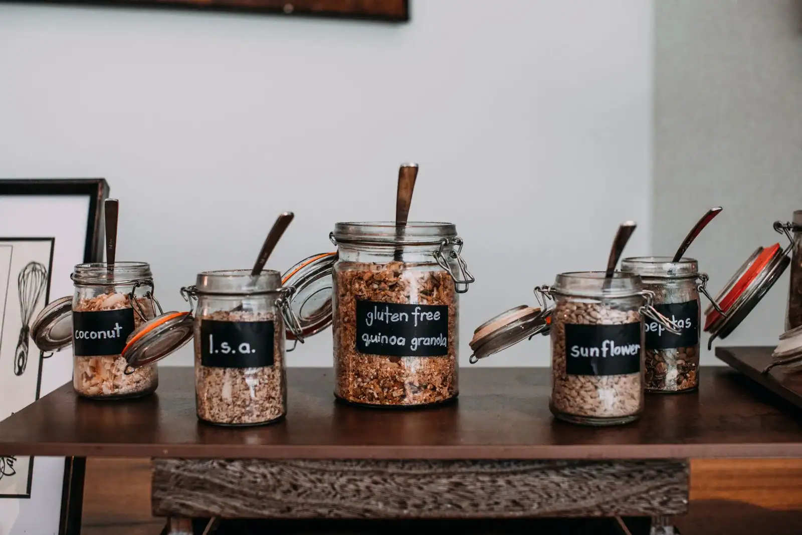 Bocaux d'ingrédients sans gluten étiquetés — granola de quinoa, graines et noix de coco — sur une table en bois