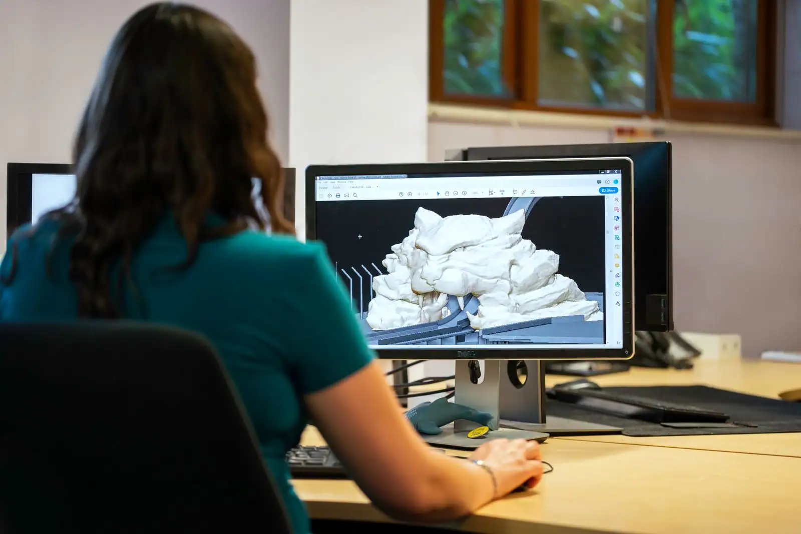Une personne travaille sur un modèle 3D affiché à l'écran, dans un bureau à deux moniteurs.