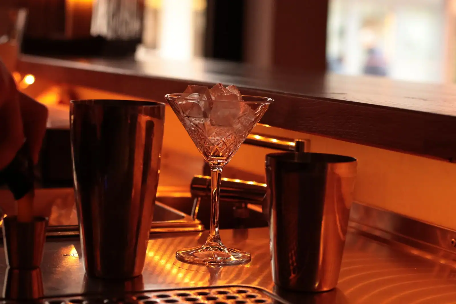 Verre à cocktail rempli de glaçons entre deux timbales à shaker, sur un comptoir de bar en inox