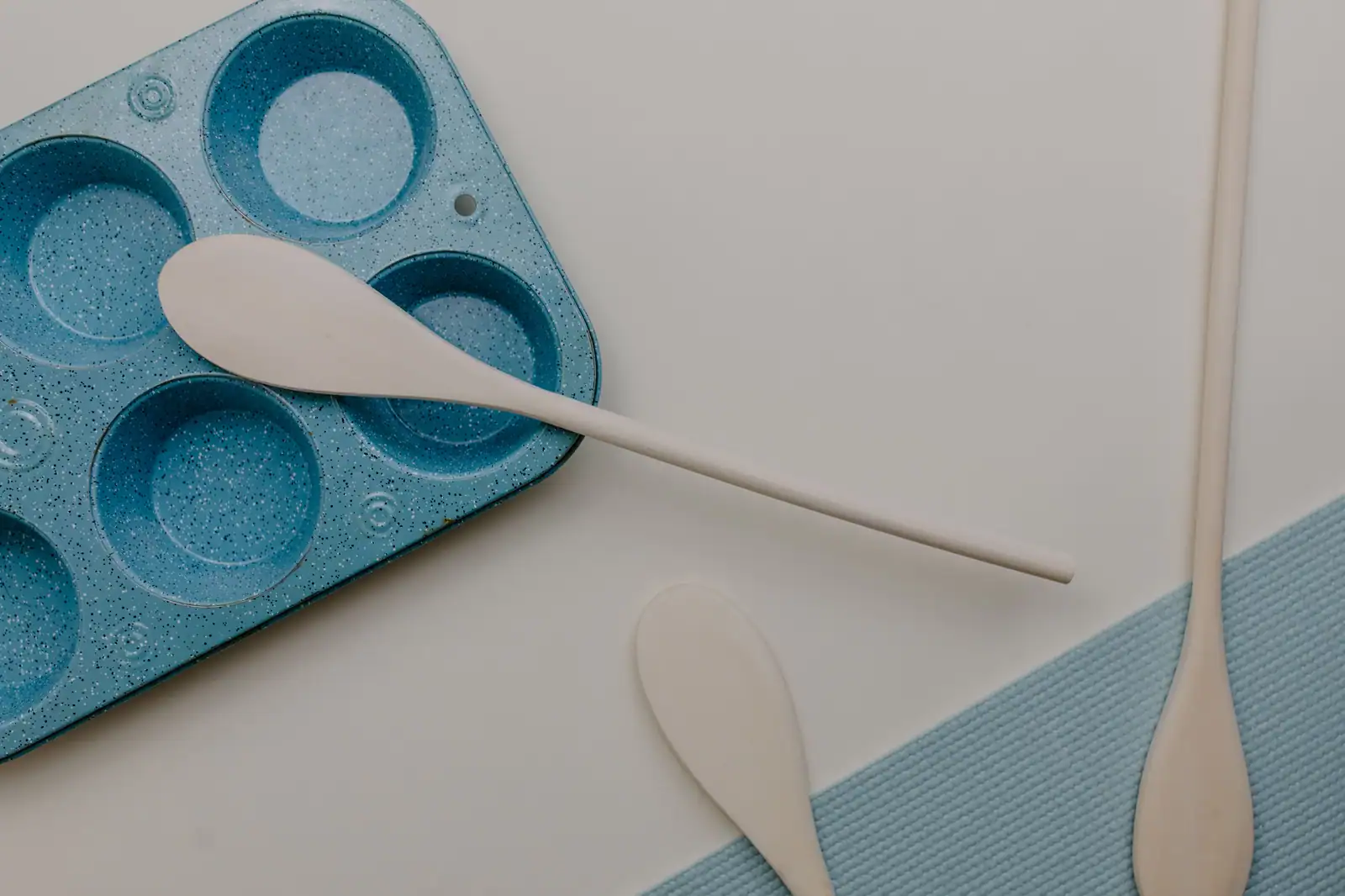 Moule à muffins bleu et trois cuillères en bois disposés à plat sur une surface claire et épurée