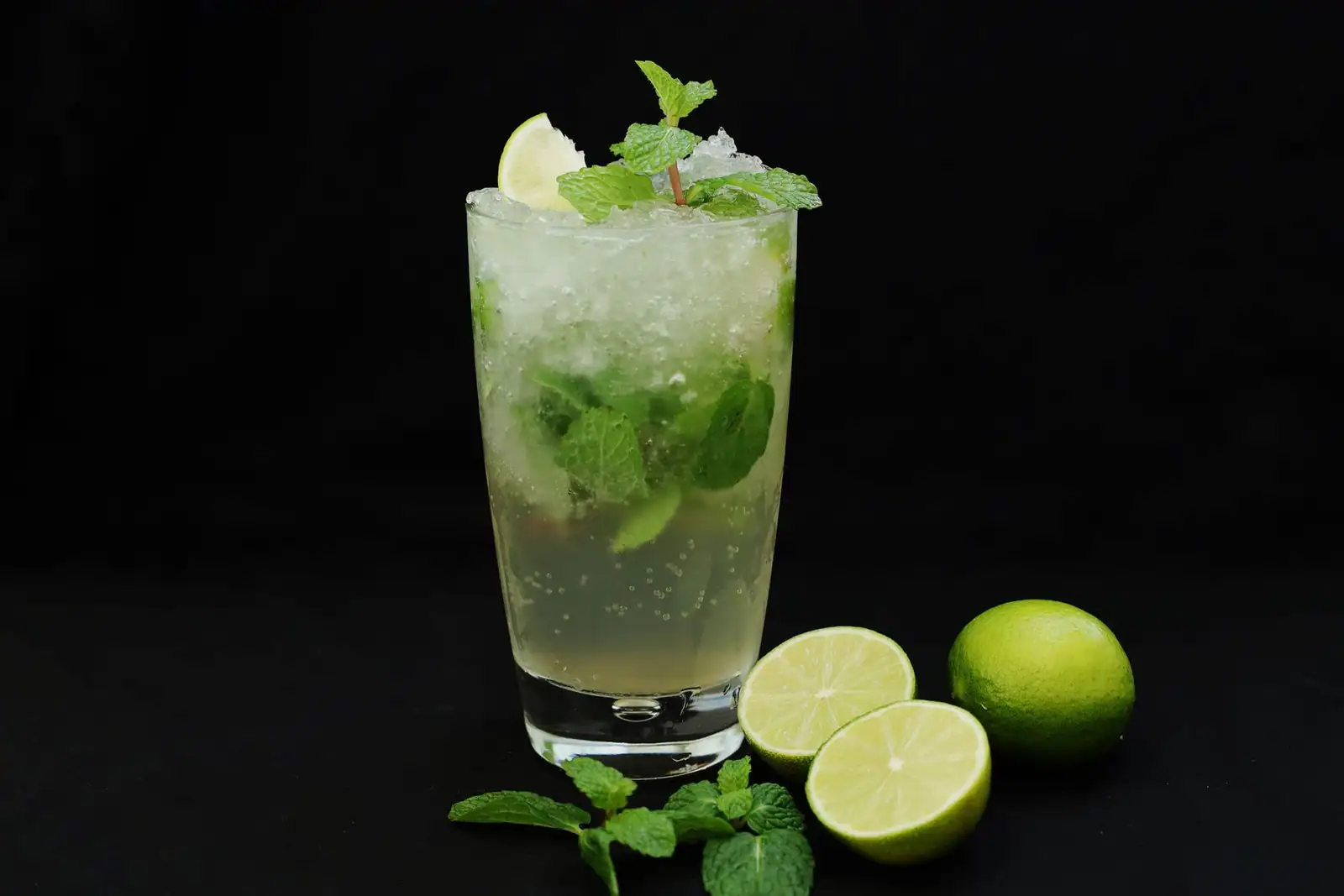 Verre droit rempli de glace pilée, de menthe fraîche et de citron vert, entouré de limes coupées en deux